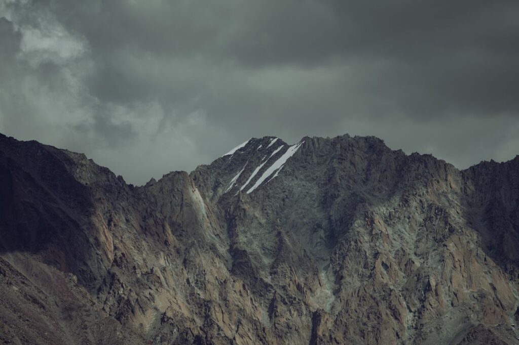 A majestic mountain range under a dramatic gray sky with rugged peaks and snow patches.